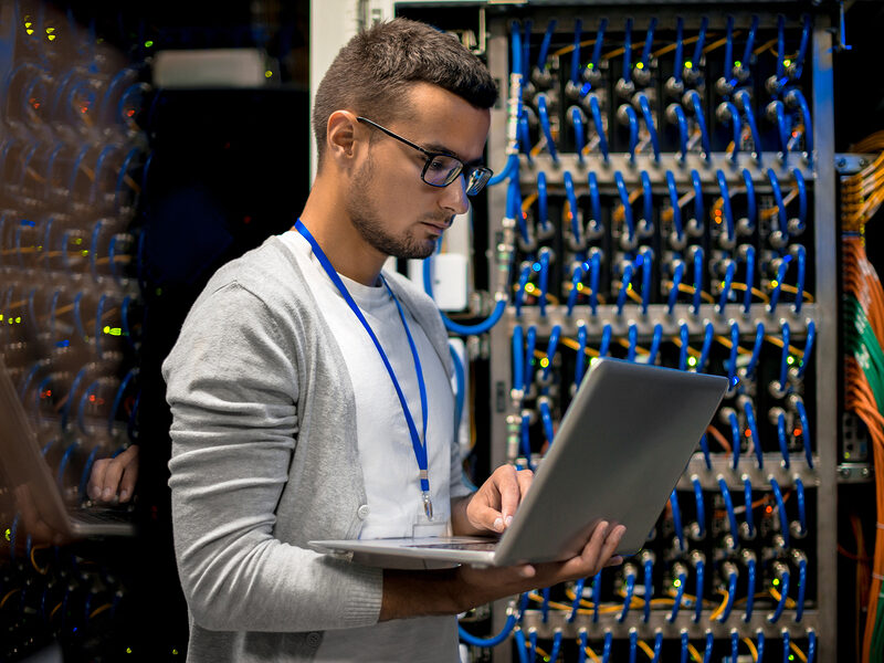 Side view  portrait of young man with laptop standing by server cabinet while working with supercomputer